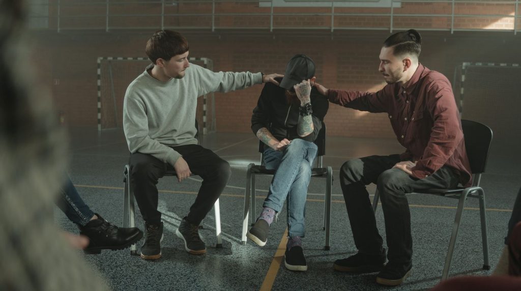 Three men sit together in a group therapy circle, one resting a hand on another man shoulder.