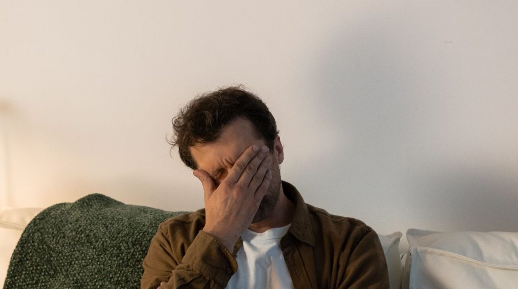 A man sits on a couch with one hand over his face, quiet and withdrawn.
