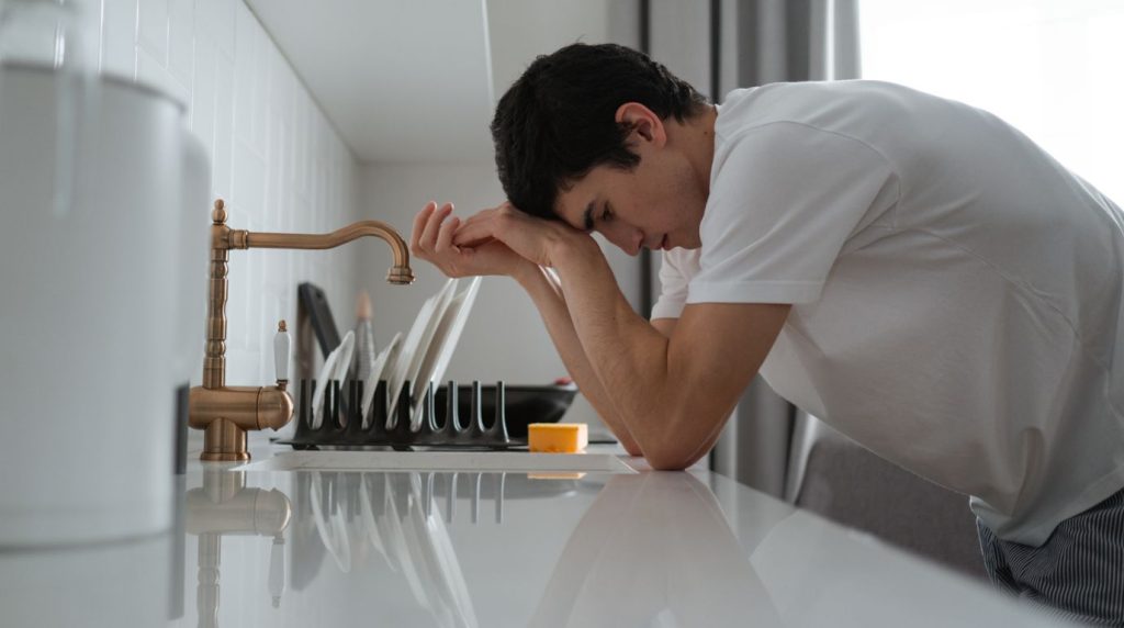A man leans on a kitchen counter with his head down, worn out in flat daylight.