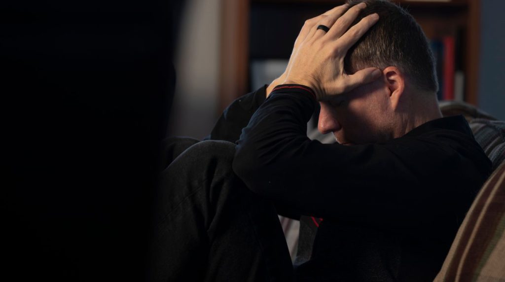 A man sits alone in a dim room with both hands in his hair, head lowered.