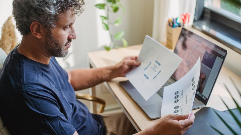 A man reviews insurance paperwork beside a laptop at a table in morning light.