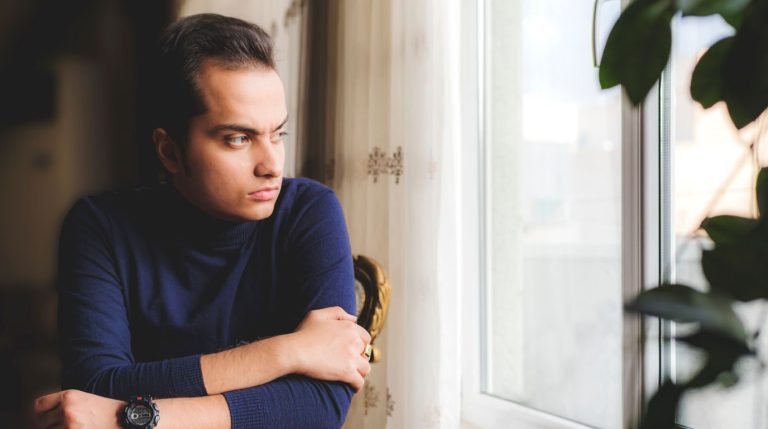 A man sits by a window in a quiet room, looking out, subdued in soft daylight.