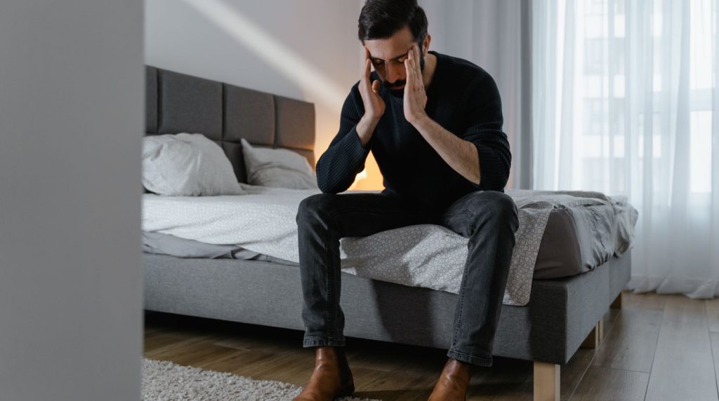 A man sits on the edge of his bed in the morning, one hand to his face, tired and unfocused.